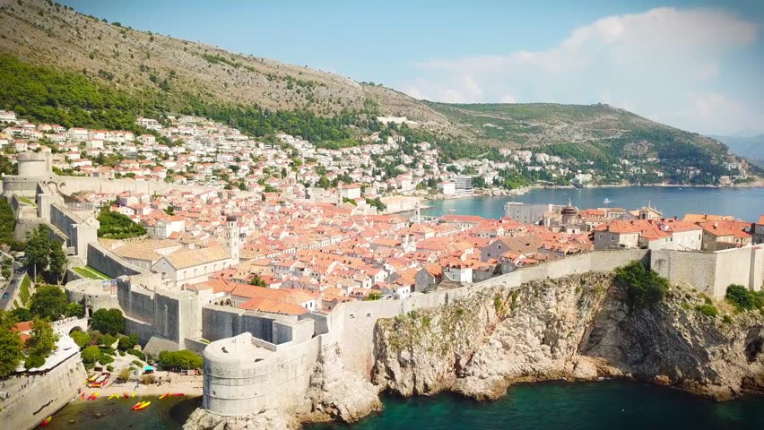 A smooth backward-moving drone shot reveals the stunning medieval walls and terracotta rooftops of Dubrovnik Old Town, Croatia, surrounded by crystal-blue Adriatic waters and dramatic coastal cliffs.