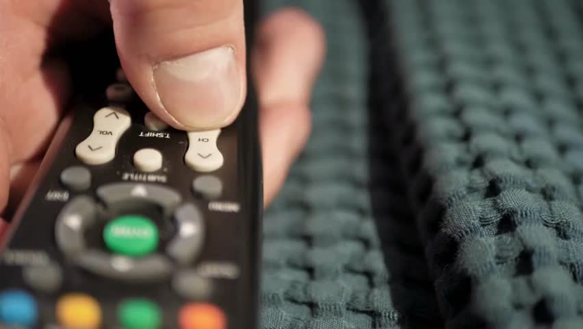 A Person Sitting on sofa while flipping through TV channels with remote control in hand. A hand controlling TV and movie channels. Close up shot of a remote control in a man's hand.