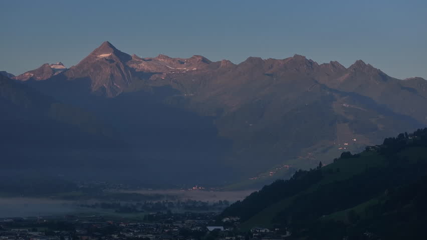 alpenglow kisses jagged mountain summits with warm pink light while valley remains cool and shadowed, cinematic aerial shot