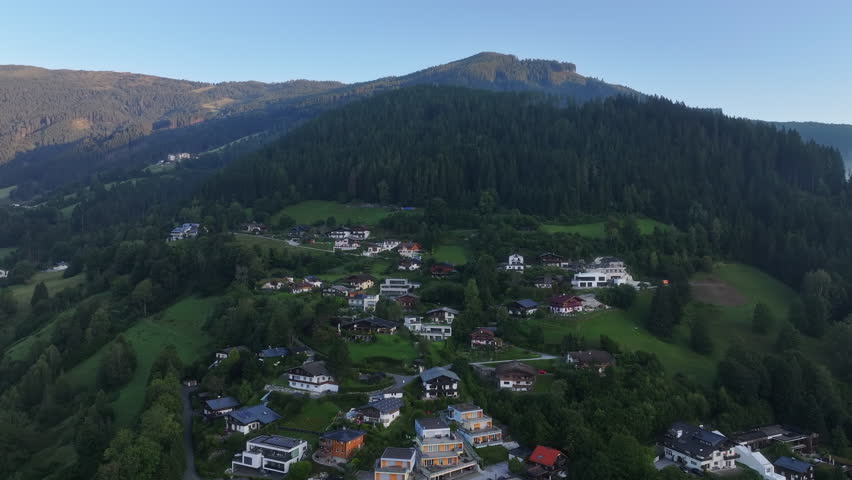 sunlit hillside village beneath dense conifer forest and high ridge, drone perspective highlights rooftops, winding roads,