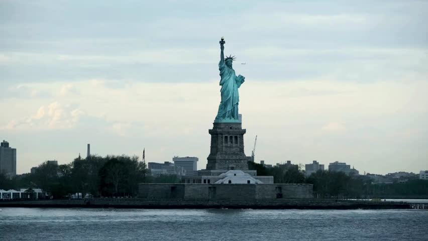 Iconic Statue of Liberty stands tall against twilight sky, surrounded by calm waters. Skyline of New York City provides striking backdrop, showcasing urban landscape.