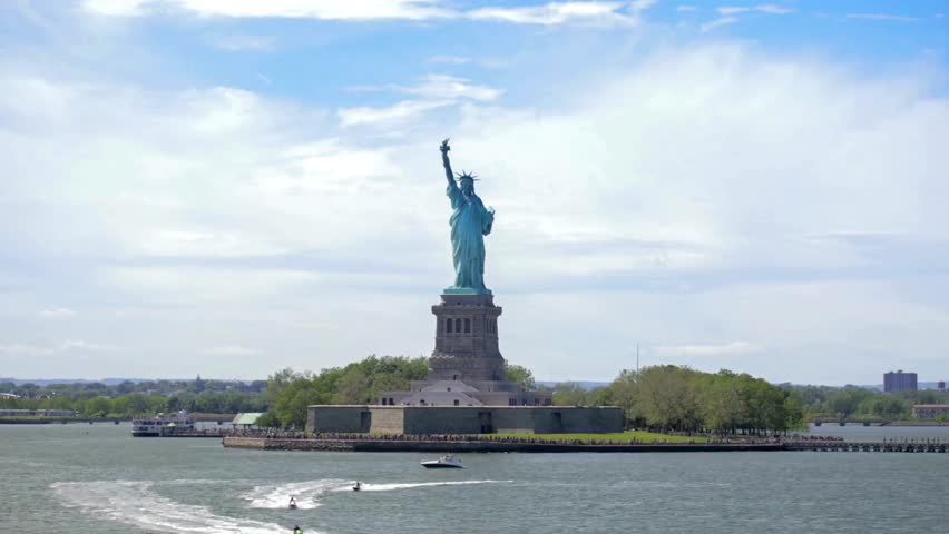 Statue of Liberty stands tall on Liberty Island. Boat moves in water near shore. Blue sky with clouds on bright day. New York City visible in background.