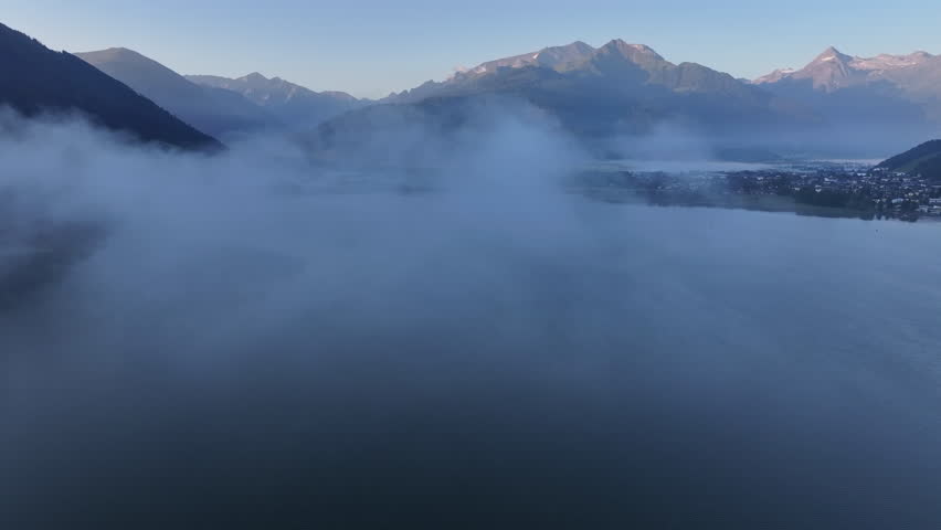dense fog filling alpine basin and lake at dawn, aerial shot shows swirling mist, hidden shoreline and distant sunlit peaks