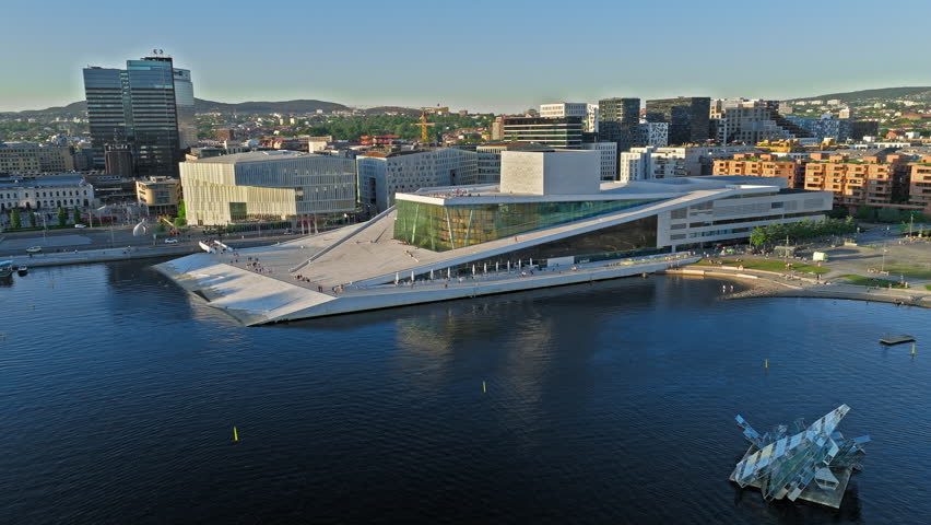 High tilt-reveal drone shot of Oslo Opera House and surrounding waterfront. Bright summer tourism aerial showing fjord, harbor boats, architecture and active sightseeing mood.