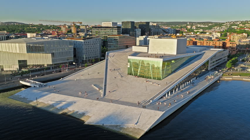 Wide drone orbit of Oslo Opera House by Oslofjord Norway. Summer daylight travel lifestyle view capturing sloped marble rooftop, people, water texture and urban scene.