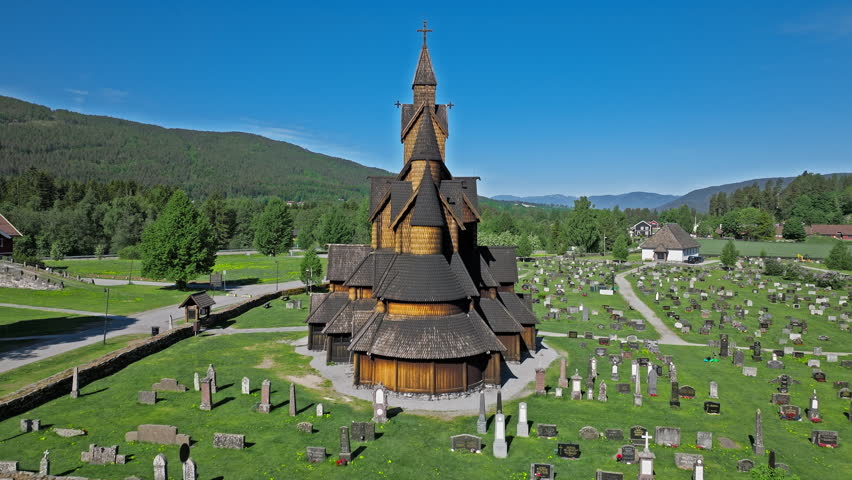 Aerial view of Heddal Stave Church in Notodden Norway showing medieval wooden towers and cemetery with grave markers. Sunny summer landscape with forests and mountains framing historic Nordic architecture for tourism travel lifestyle.