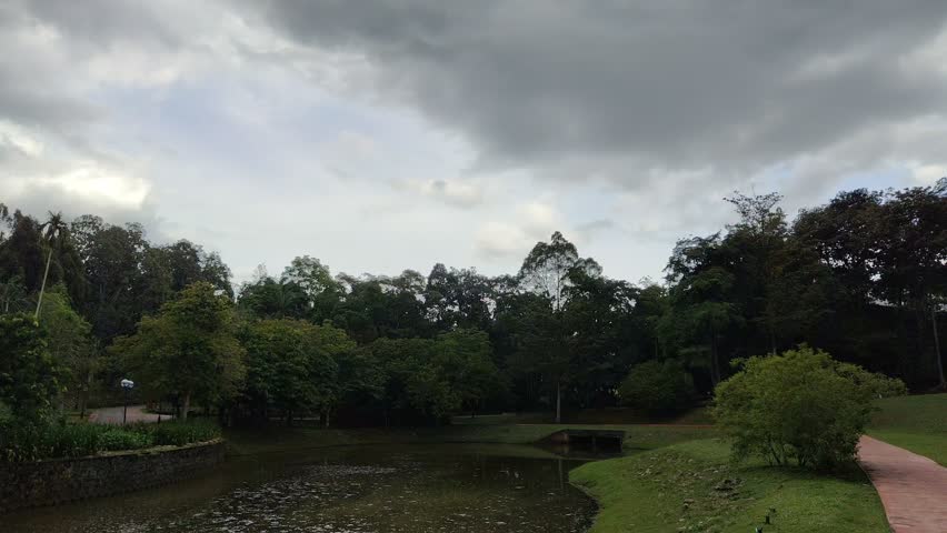 ​A tranquil park pond surrounded by lush tropical greenery under a heavy, dark, and dramatic overcast sky. Paved walkway on the grassy bank