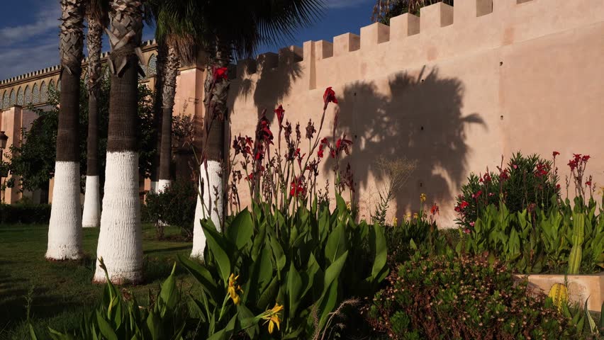 A public garden with yellow and red flowers in front of a rampart. On the left side, there are a few palm trees.