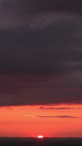 Vertical screen sunset over horizon with crimson sky dark storm clouds roll above small sun disk dipping behind appalachian ridge, deep red orange band shimmer across distant landscape, cinematic mood