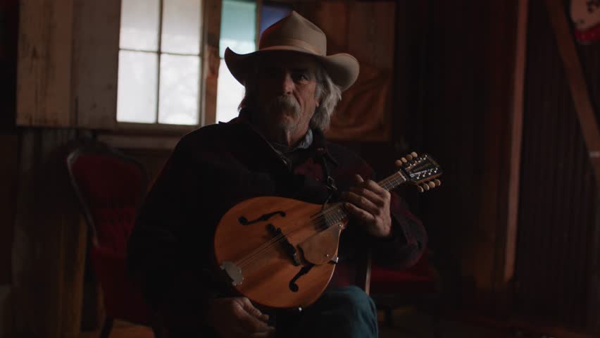 Man With Mustache Wearing Cowboy Hat Holding Mandolin String Instrument Inside of Shadowy Saloon