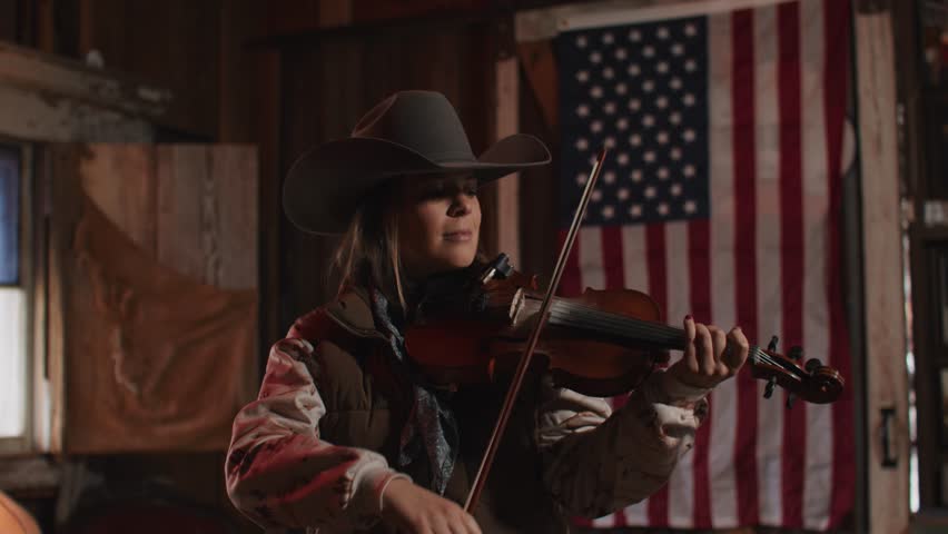 Woman Wearing Cowboy Hat and Vest Playing Fiddle Violin Music In Front of American Flag Inside of Old Rustic Saloon