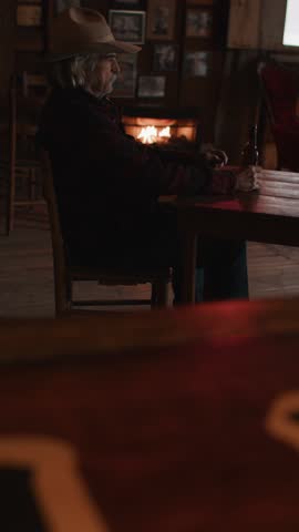 Man With Mustache Wearing Red Flannel and Cowboy Hat Sitting With Beer Bottle in Front of Fireplace in Old Rustic Saloon