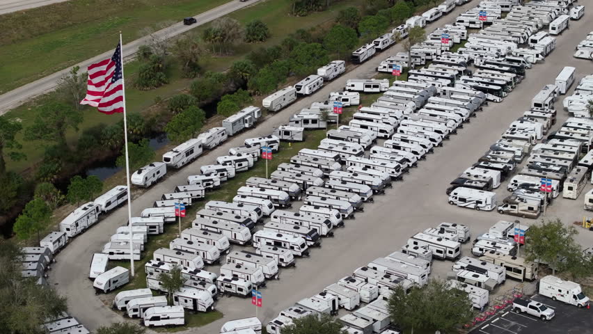 Large parking lot of local dealer with many new RV trailers parked for sale. Distribution of manufactured camping vehicles.
