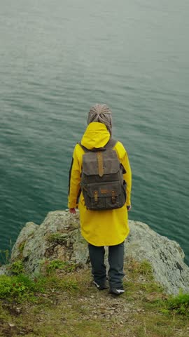 A person in a vibrant yellow raincoat standing at the edge of a scenic cliff, taking in the breathtaking view of the calm waters and mountains beyond, capturing the moment with a smartphone.