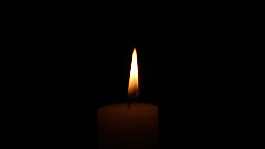 A single candle burns in a dark indoor setting as its flame flickers gently before rapidly dimming and disappearing, leaving the scene in complete darkness with subtle fading movement.