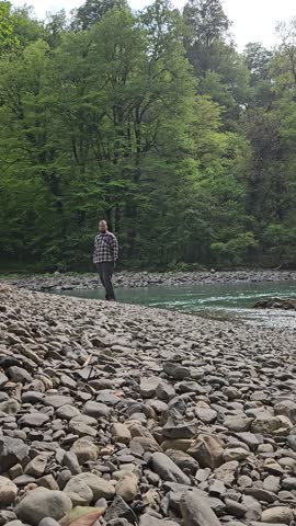 Man standing on a rocky shore next to a river. He is wearing a plaid shirt and a hat