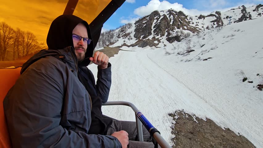 Man in a black jacket sits in a chair looking out over a snowy mountain. He is wearing a black hoodie and glasses