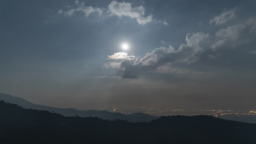 A bright full moon shines over a distant city in a dark valley, framed by rolling mountain ridges in Asia. Stunning night scene captured during a cold twilight for travel and celestial photography