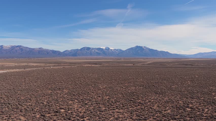 Aerial view moving across the arid sagebrush plains of Northern New Mexico toward the majestic Sangre de Cristo Mountains. Captures the distinct high desert terrain.