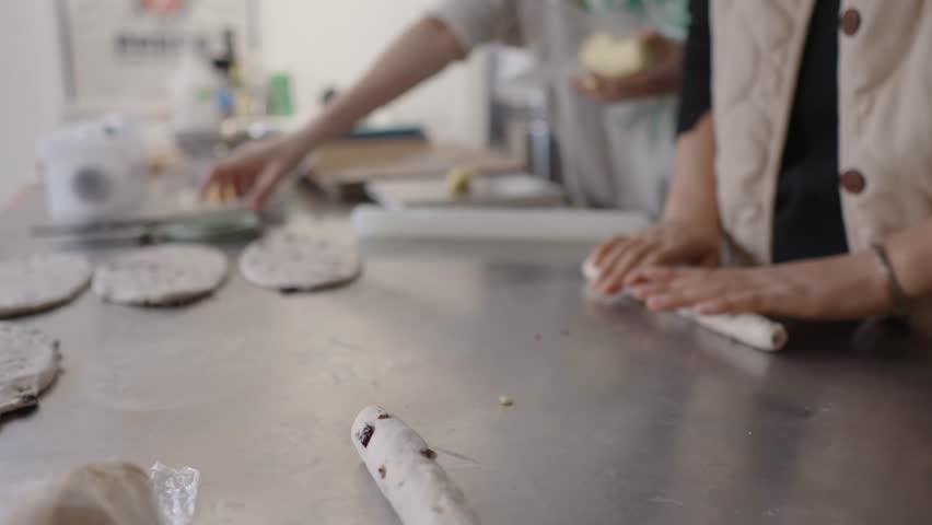 Close up view of baker hands skillfully rolling log of fresh raisin dough on stainless steel table in commercial kitchen, preparing it for baking in slow motion shot, enjoying cooking in the morning