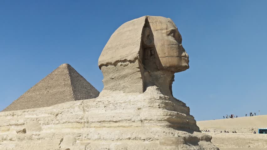 Great Sphinx of Giza, carved from limestone bedrock, faces right, with its head and body clearly visible against a bright blue sky. The Pyramid of Khufu rises in the background.