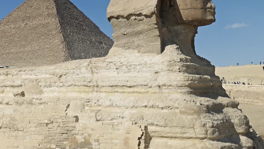 Great Sphinx of Giza, carved from limestone bedrock, faces right, with its head and body clearly visible against a bright blue sky. The Pyramid of Khufu rises in the background.