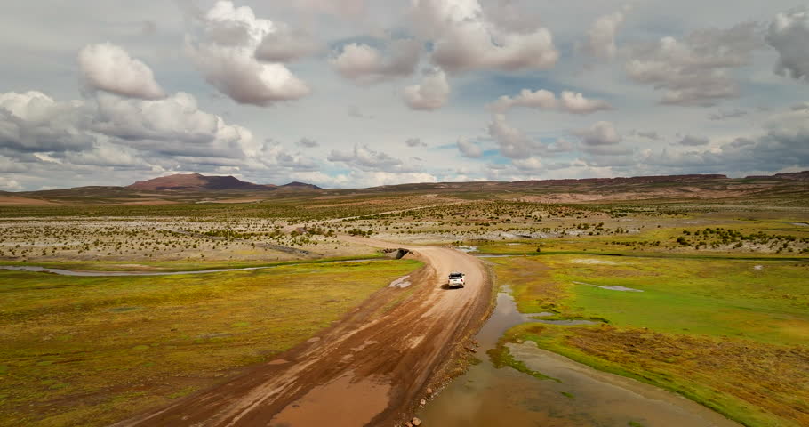 Aerial drone view of a white pickup truck driving on a dirt road in the Siloli Desert, Bolivia. The vehicle navigates the arid high-altitude landscape with mountains under a cloudy sky