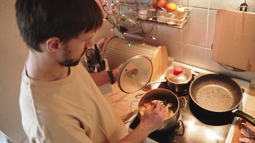 Man opens lid of pot of soup on stove and stirs it with spoon, overhead shot. Male person cooks hot entree with meat in home kitchen, warm light.