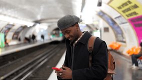 Millennial black man smiling while using his smartphone and texting on a subway platform as a train arrives. Young commuter waits for public transportation while connected to the internet - Powered by Shutterstock - Get 15% off with code: PIKWIZARD15