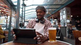 Stylish young black man wearing a beret sitting at a table in a pub, browsing on a digital tablet while enjoying a pint of beer, showcasing a modern, connected, and relaxed lifestyle - Powered by Shutterstock - Get 15% off with code: PIKWIZARD15
