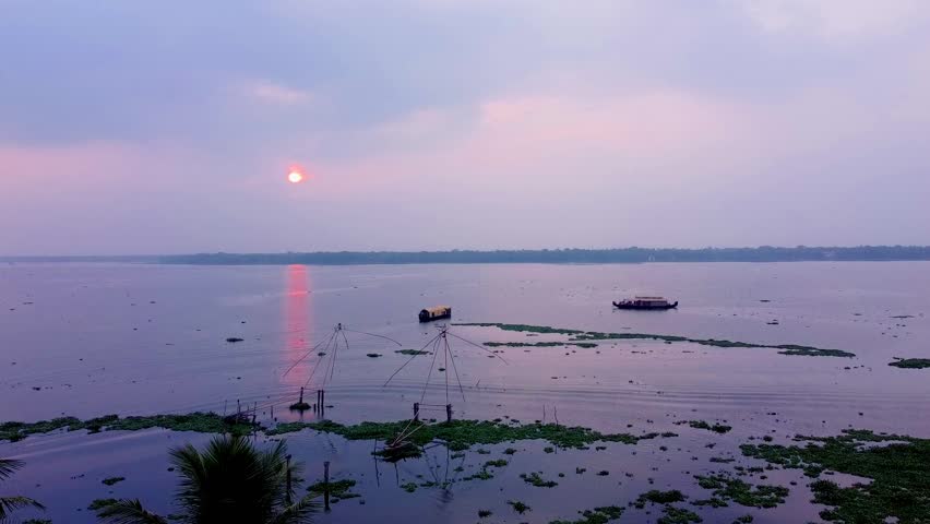 Wide aerial of Chinese fishing nets and houseboats at sunset over Kerala backwaters, with a hazy purple sky and sun reflection, showing the vast lake.