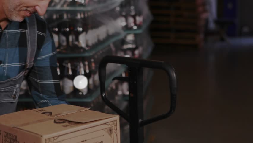 Adult male logistics worker inspecting box, pushing jack, smiling, showing network icons expanding. Warehouse, pallet, inventory, cardboard, aisle, industrial, connectivity