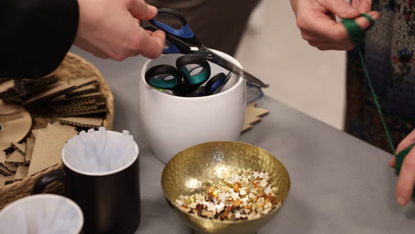 A close-up image showing a person picking golden and wooden star-shaped confetti from a decorative brass bowl on a crafting table. Scissors, glue sticks, and cardboard shapes are visible nearby, indic