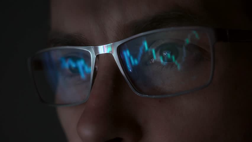 Face of trader wearing eyeglasses with reflection cryptocurrency charts. Close-up portrait of man analysing stock market. 
