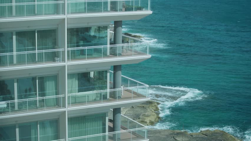 Right pan of modern seafront apartment balconies overlooking turquoise Mediterranean water in Malta, waves crashing on rocky shore below under bright daylight.