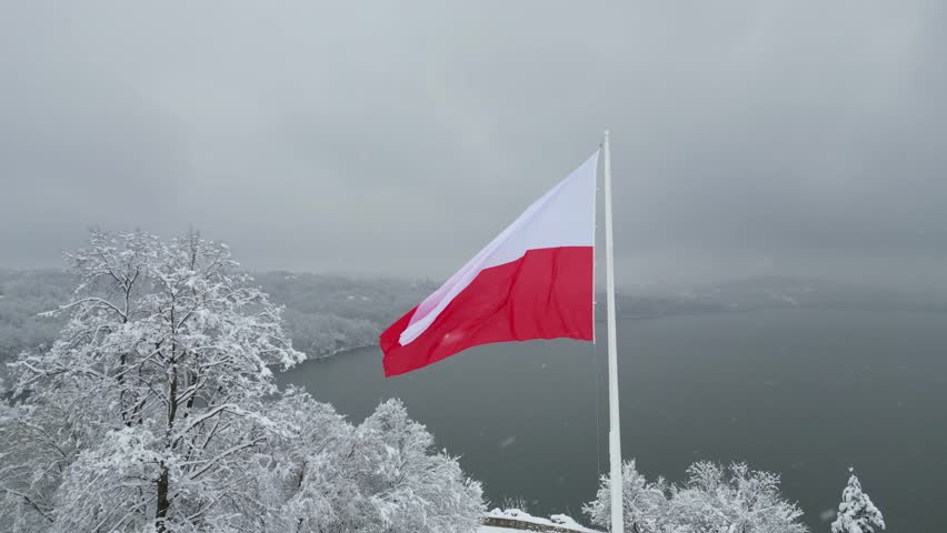 Polish flag gracefully flutters in the cold winter breeze against a serene lake and snow-covered forest. Flag of Poland waves gently in the winter wind with tranquil lake and snowy trees at background