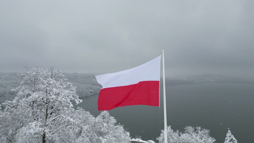 Polish flag gracefully flutters in the cold winter breeze against a serene lake and snow-covered forest. Flag of Poland waves gently in the winter wind with tranquil lake and snowy trees at background