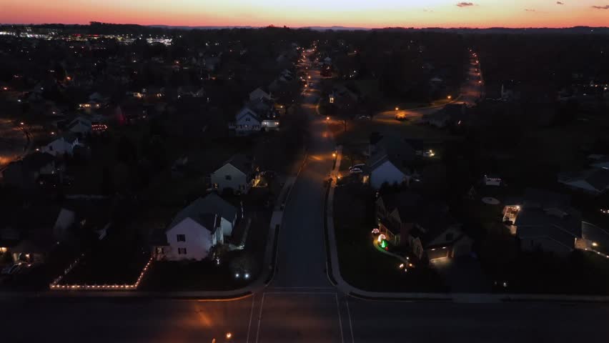 Slow flight over illuminated street in sub urban housing area of american town. Dusk sunset scene in fall. Single family houses and homes in USA. Aerial Birds Eye shot.
