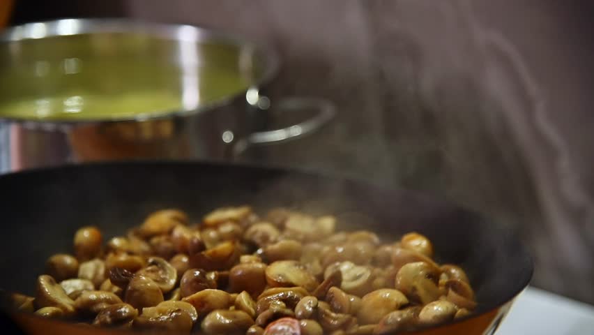 A slow-motion close-up of sliced small porcini mushrooms being fried in a frying pan in a home kitchen. Stirring the mushrooms as they fry by tossing them in the pan.