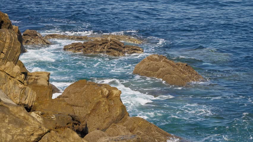 Clear blue ocean water washes over textured, sun-drenched coastal rock formations.