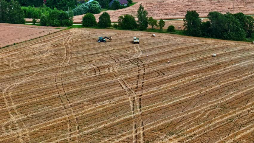 Tractor with front loader lifting hay bales onto trailer in harvested crop field