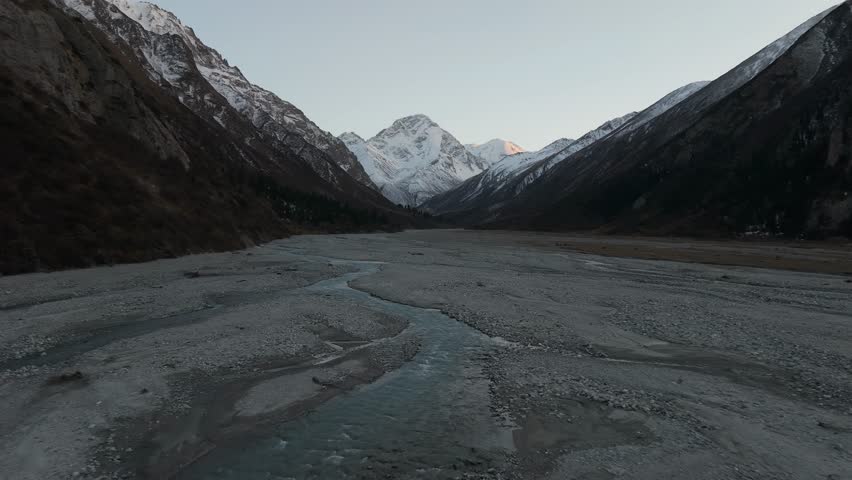 Majestic mountain landscape reveals a flowing river winding through rocky terrain, showcasing snow-capped peaks and serene ambiance, as the camera pans across the breathtaking scenery