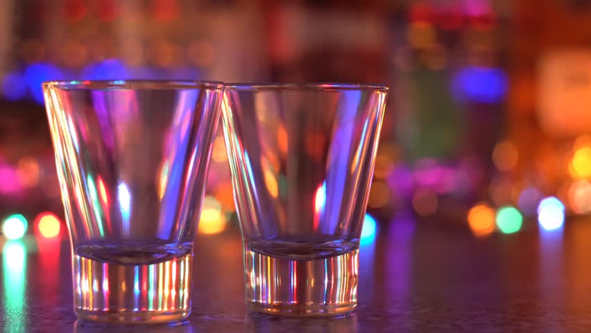 Clear alcoholic beverage is poured into two shot glasses on a reflective surface, illuminated by colorful, blurred lights, evoking a festive and lively party atmosphere