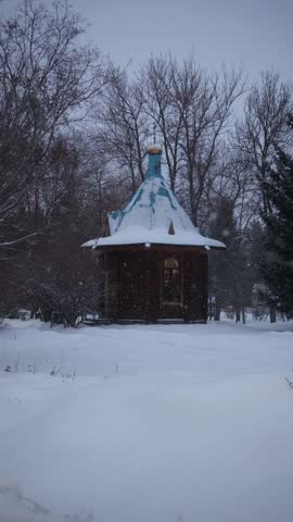 A wooden chapel standing in a quiet park as snow gently falls, creating a serene and atmospheric winter scene. The soft flakes, warm natural textures of the wood, and peaceful surroundings evoke a sen