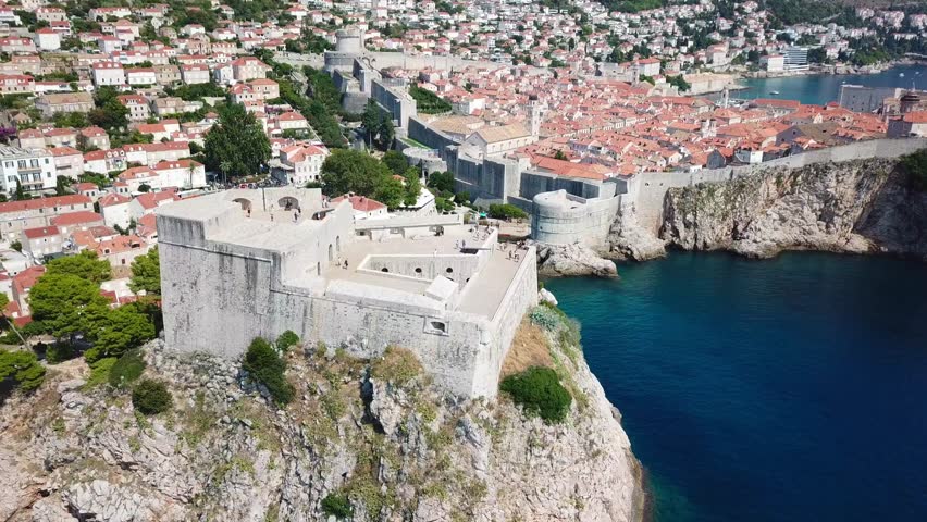 Aerial drone orbit circling the massive Fort Lovrijenac in Dubrovnik, Croatia, revealing dramatic views of the rocky cliffs, the Adriatic Sea, and the historic Old Town walls.