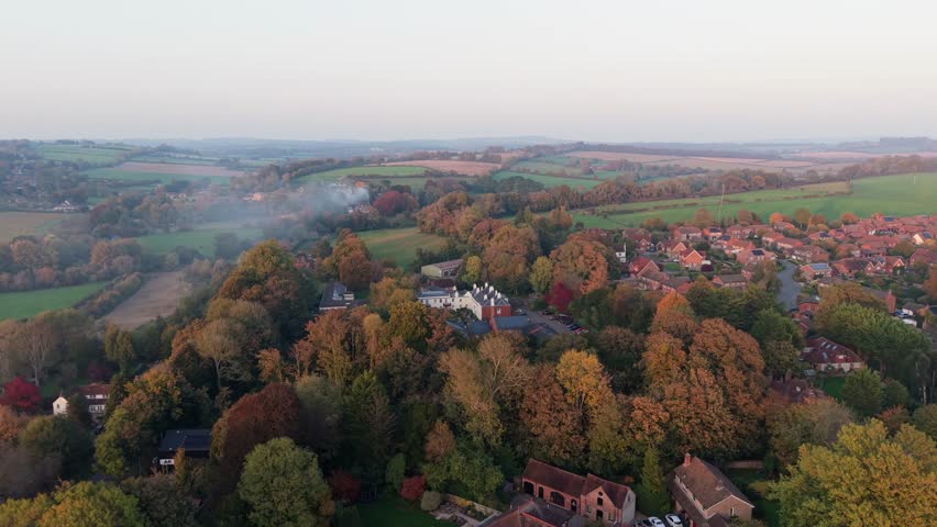 Aerial drone footage flying backwards over a historic english village. An old church and red-roofed houses nestled among autumnal trees at sunset