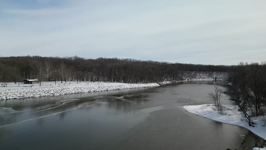 Slowly rising up over the Iowa River in the winter, just north of Iowa City and south the dam on the river. The water is flowing but the shore is covered in snow. 4k, drone, flying up.