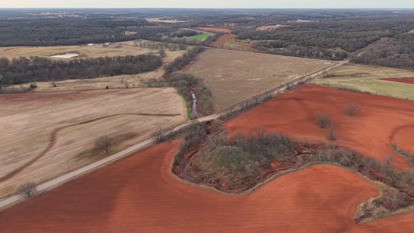 Aerial drone view of red dirt Oklahoma farmland with a winding creek flowing through winter agricultural fields. Bare trees and patchwork farmland surround the waterway, showing earthy tones and seasonal textures across a quiet rural landscape.