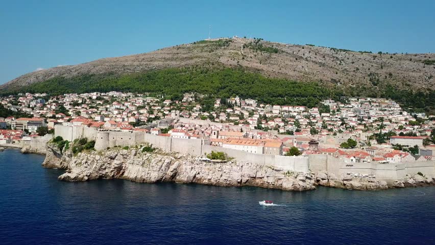 A stunning drone shot showcasing the sea-facing walls of Dubrovnik, Croatia, perched on cliffs with the imposing Mount Srđ rising in the background.