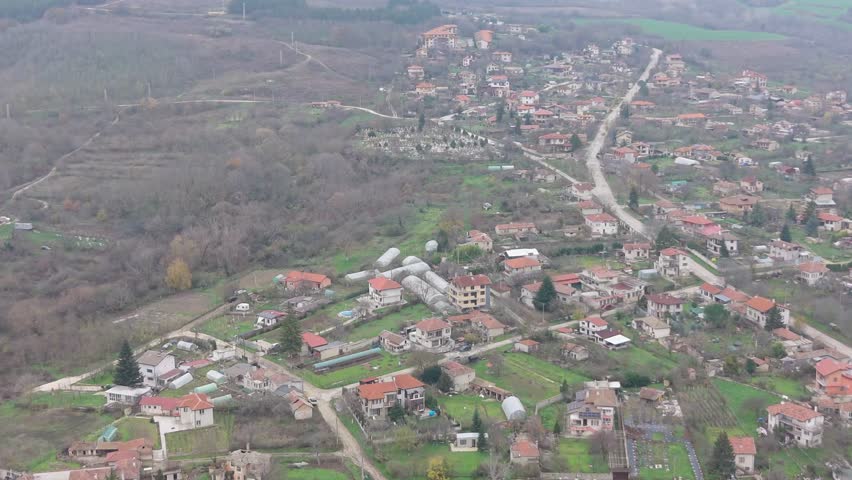 Aerial drone view of a small rural village surrounded by hills and fields, capturing rooftops, winding roads, and peaceful countryside scenery.